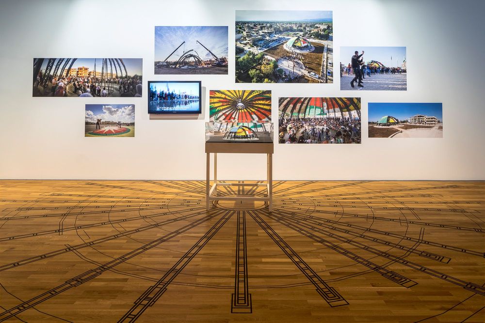 Die Ausstellunngsansicht zeigt einen kleinen Tisch in der Mitte der Aufnahme, auf dem das Modell einer Kuppel steht. Dahinter an der Wand hängen verschiedene Fotografien. Am Boden unter der Kuppel ist davon ausgehend eine Skizze angebracht.