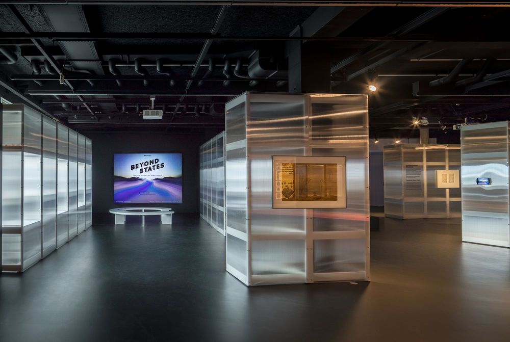 Die Ausstellungsansicht zeigt mehrere aufgebaute Ausstellungswände, die leicht transparent sind. An der hinteren Wand projiziert ein Beamer ein Bild gegen eine Leinwand. Davor steht eine runde Sitzbank für Besucher*innen.