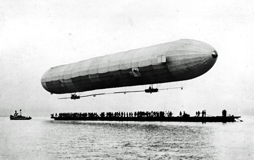 Schwarz-weiß-Fotografie eines Zeppelins, der knapp über einer Plattform auf dem Wasser schwebt. Zahlreiche Menschen stehen auf der Plattform, im Wasser daneben befindet sich ein Boot.