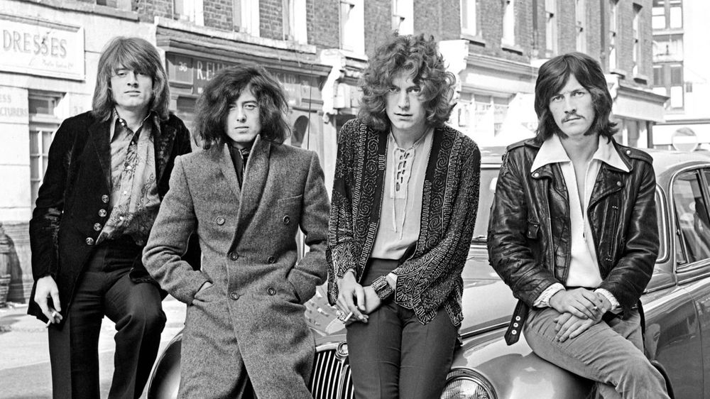 Die schwarz-weiß Fotografie zeigt die Rockband Led Zeppelin. Vier Männer mit langen Haaren sitzen oder lehnen an einem Auto und schauen in die Kamera.