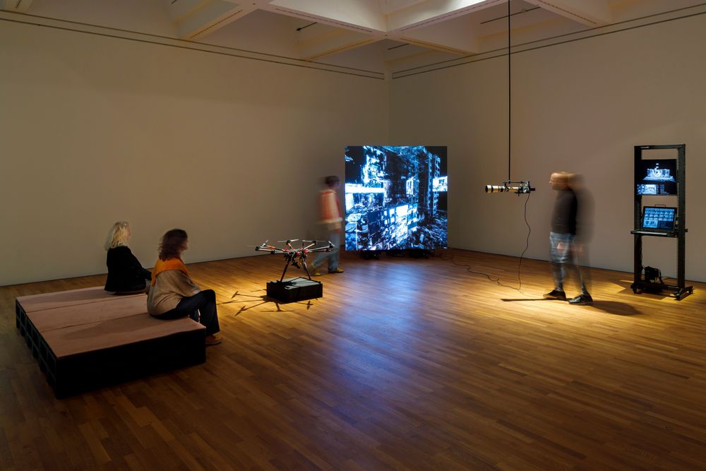 Die Ausstellungsansicht zeigt zwei Frauen die auf einer Bank sitzen und eine Drohne vor sich betrachten. Ein Mann auf der rechten Seite betrachtet eine große Kamera, die von der Decke hängt. 