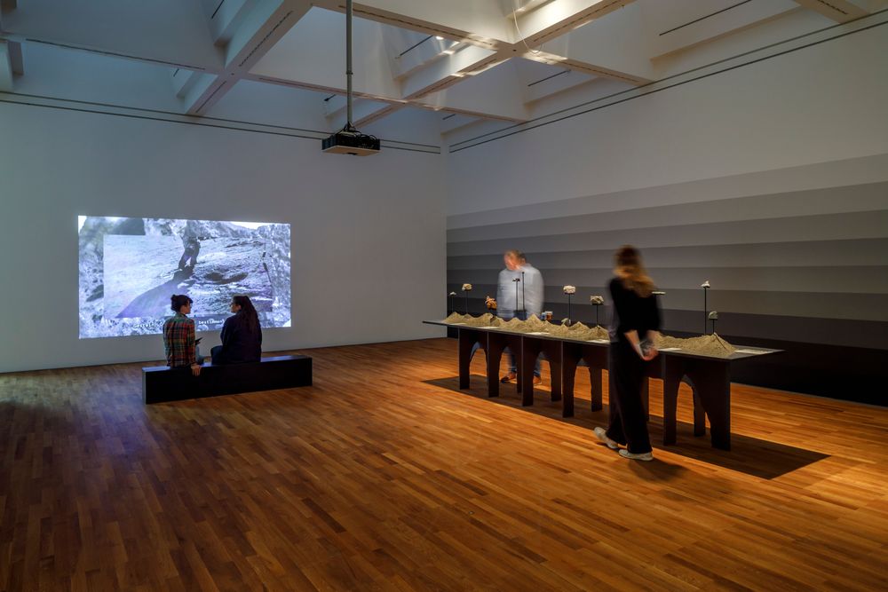 Die Ausstellungsansicht zeigt zwei Frauen die auf einer Bank sitzen und sich unterhalten. Hinter ihnen wird ein Video an eine Wand projiziert. Daneben befindet sich ein Tisch mit verschiedenen Ausstellungsobjekten darauf.