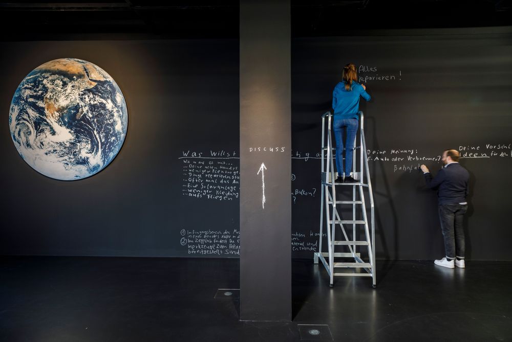 Die Ausstellungsansicht zeigt zwei Personen, die mit weißer Kreide auf eine schwarte Wand schreiben. Die Frau steht auf einer Leiter. 