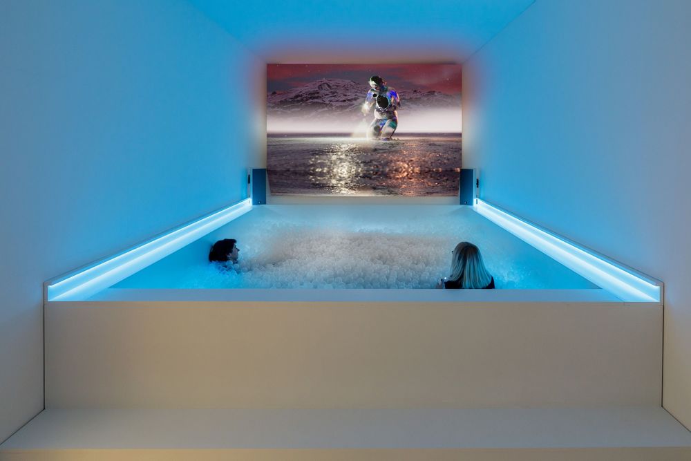 Installationsansicht auf ein weißes Bällebad, das von unten blau ausgeleuchtet ist. Zwei Personen sitzen im Bällebad. An der RAumrückwand sieht man ein Still aus dem Kunstspiel.
