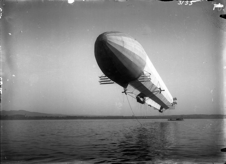 Die historische Aufnahme zeigt einen Zeppelin der über Wasser schwebt. In der Ferne dahinter ist ein Boot zu erkennen.