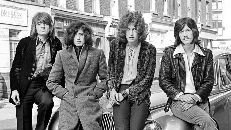 Die schwarz-weiß Fotografie zeigt die Rockband Led Zeppelin. Vier Männer mit langen Haaren sitzen oder lehnen an einem Auto und schauen in die Kamera.