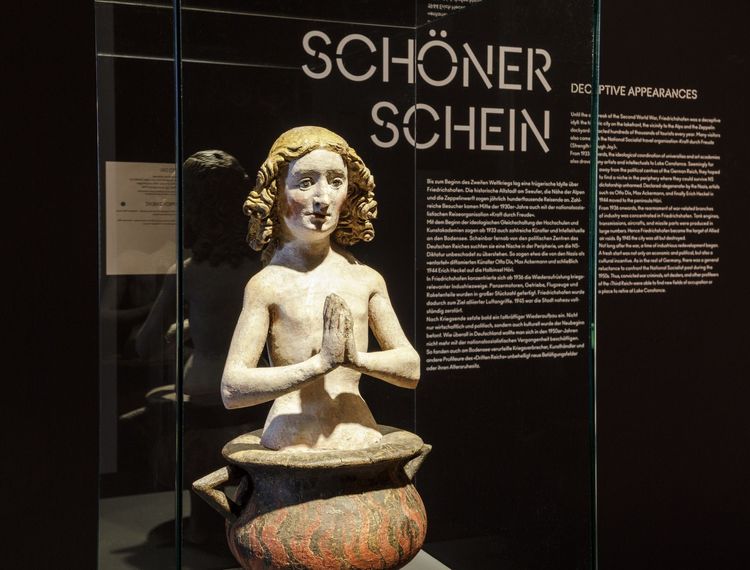 Die Aufnahme zeigt die Skulptur eines Jungen der mit betenden Händen in einem brennenden Kessel sitzt..